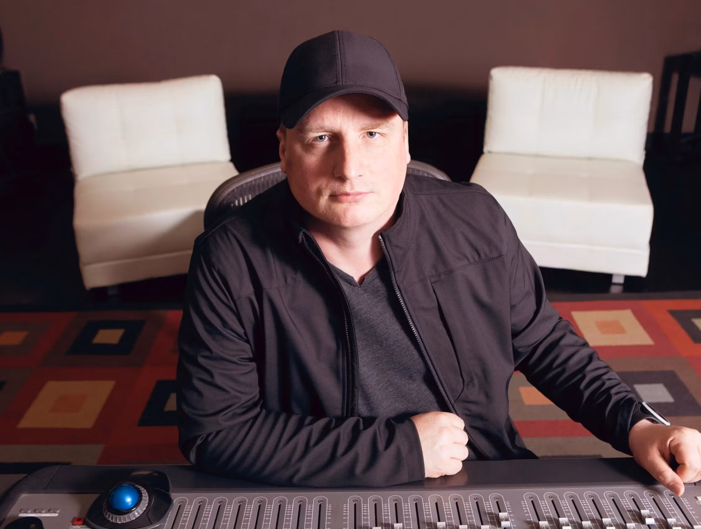 Studio portrait of Richard Furch wearing a black cap and jacket, sitting at a large mixing console with geometric patterned carpet and white chairs in the background.
