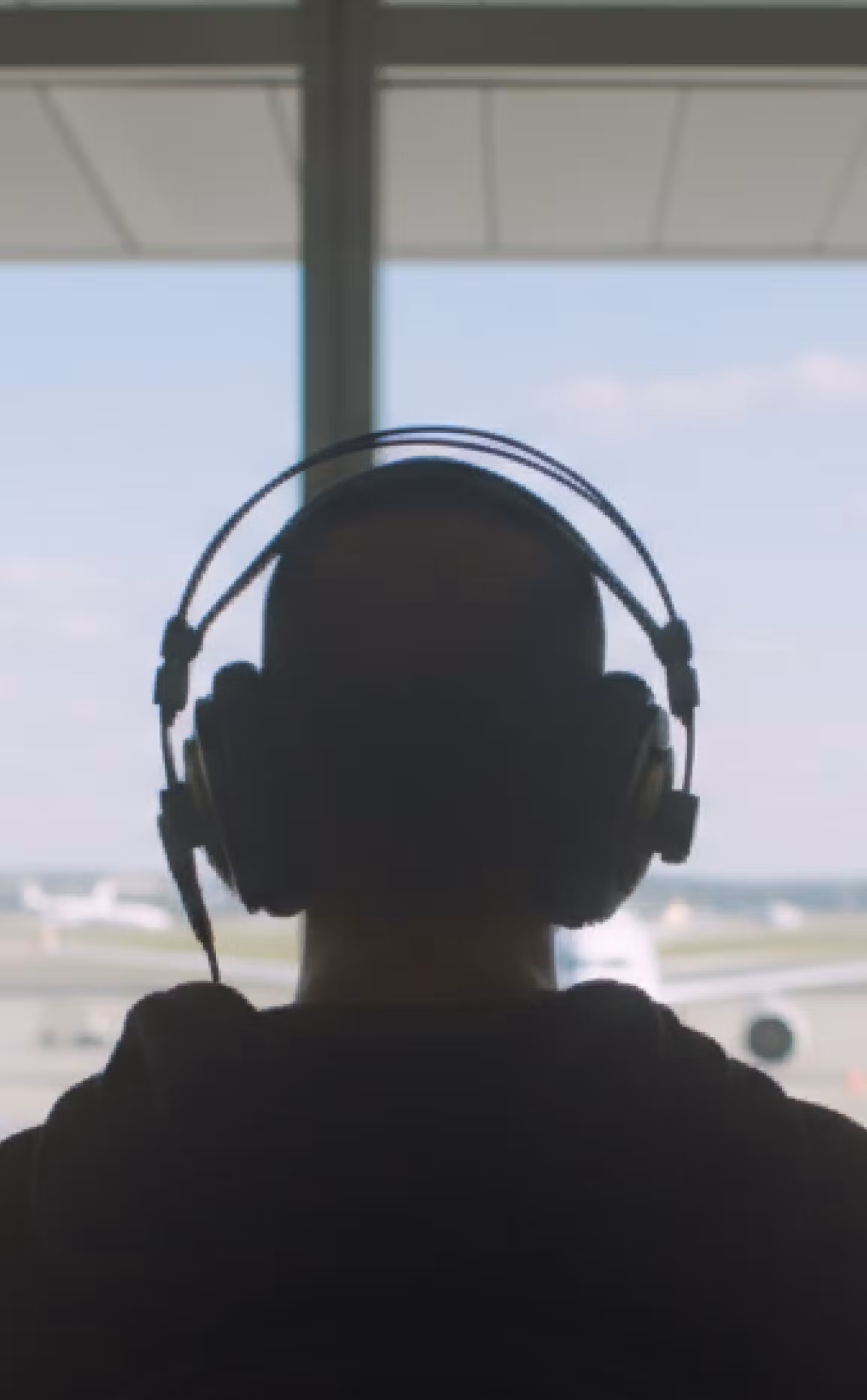Person wearing large over-ear headphones sitting by an airport terminal window, looking out at airplanes parked on the runway under daylight.