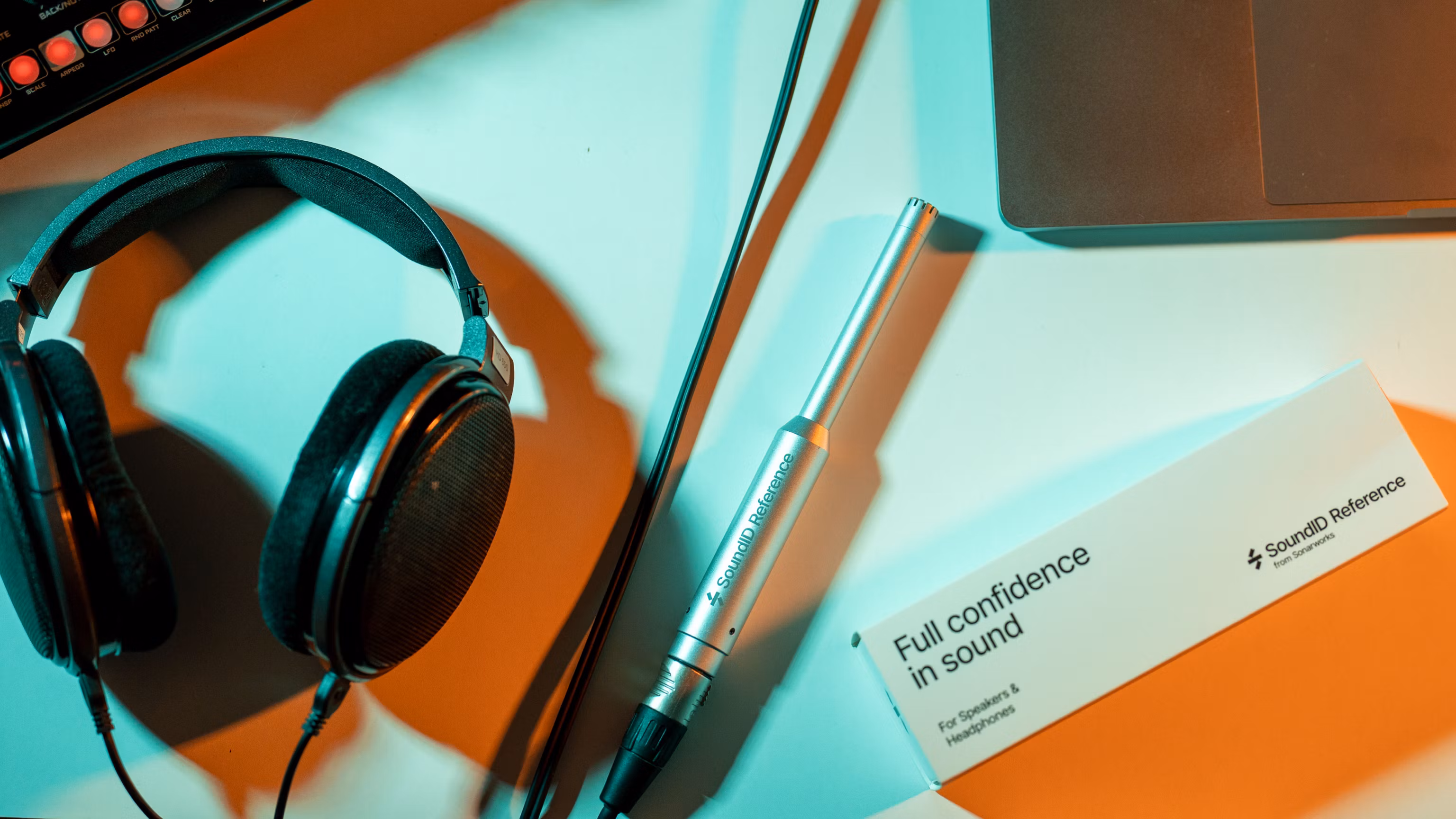 Studio desk with SoundID Reference measurement microphone, open‑back headphones, product box, and laptop under teal–orange lighting.