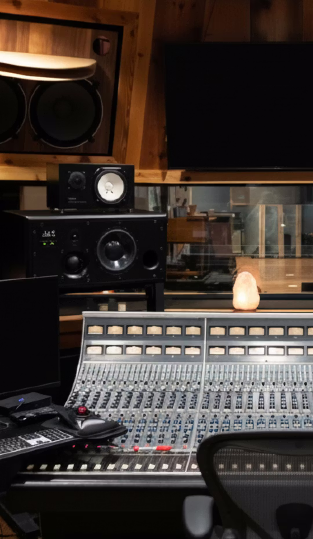 Recording studio control room with analog mixing console, stacked monitor speakers, TV screen, and wood-paneled walls.