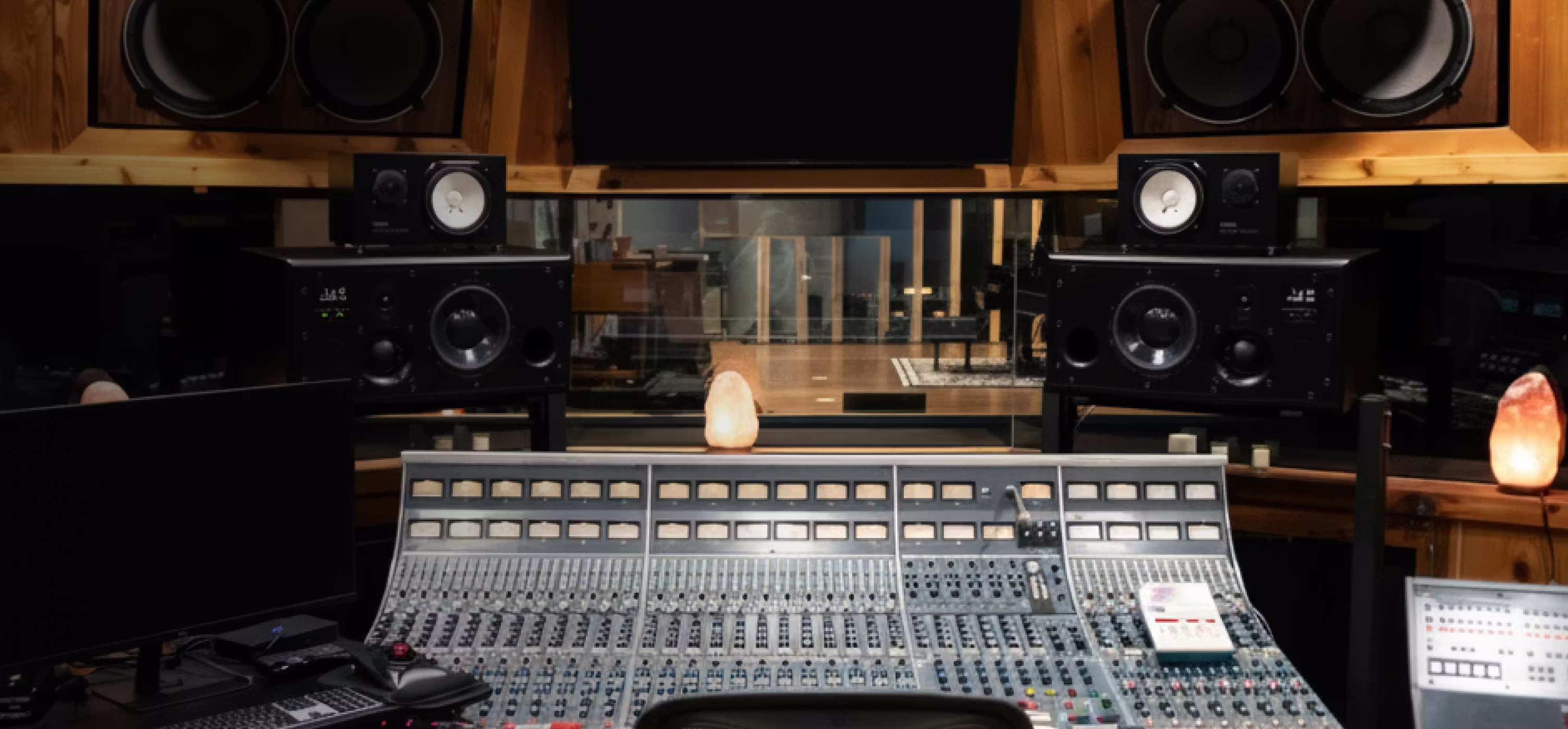 Recording studio control room with large analog mixing console, stacked monitor speakers, and wood-paneled walls behind glass.