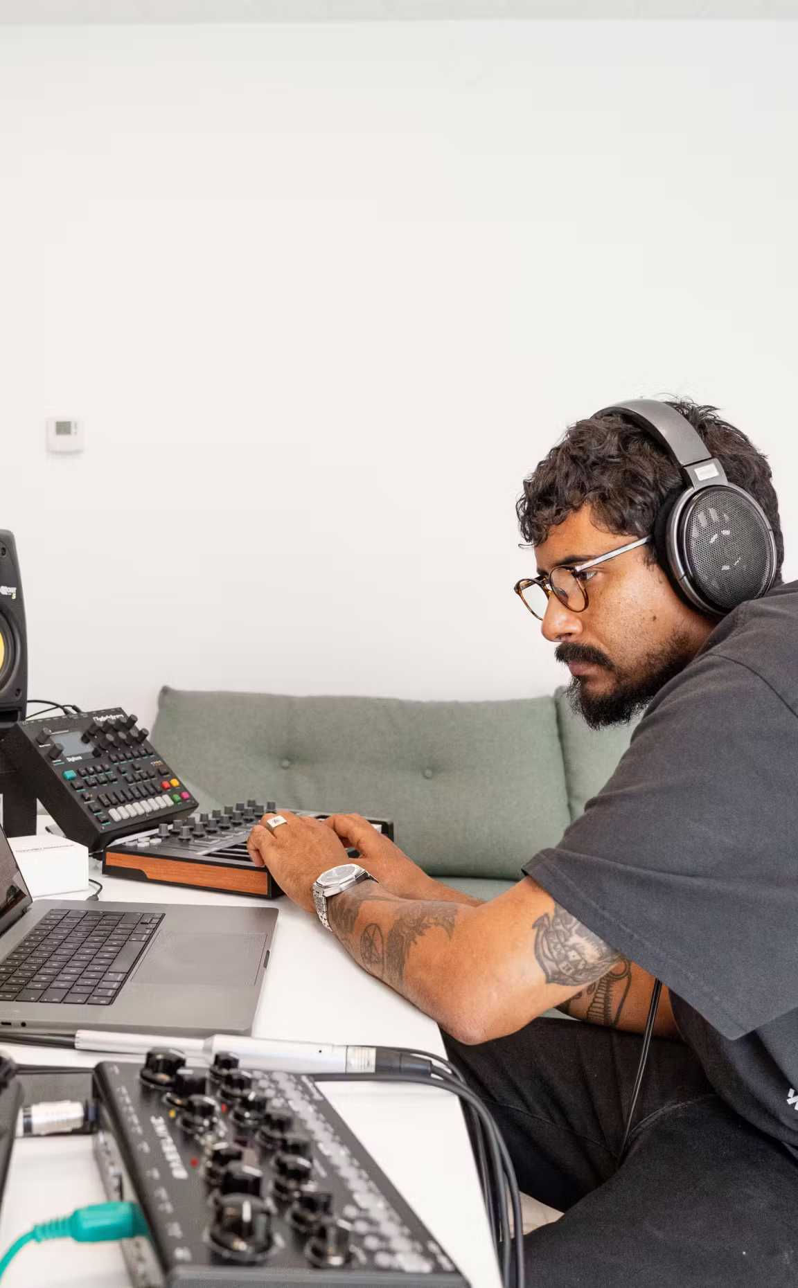 Music producer wearing open-back studio headphones working on a MIDI controller and laptop in a home studio setup with audio gear and mixing equipment.